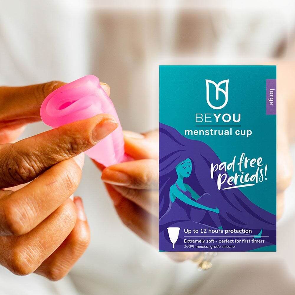 Person folding a pink BeYou menstrual cup next to product packaging that highlights 'pad free periods' and 12-hour protection for first-time users.