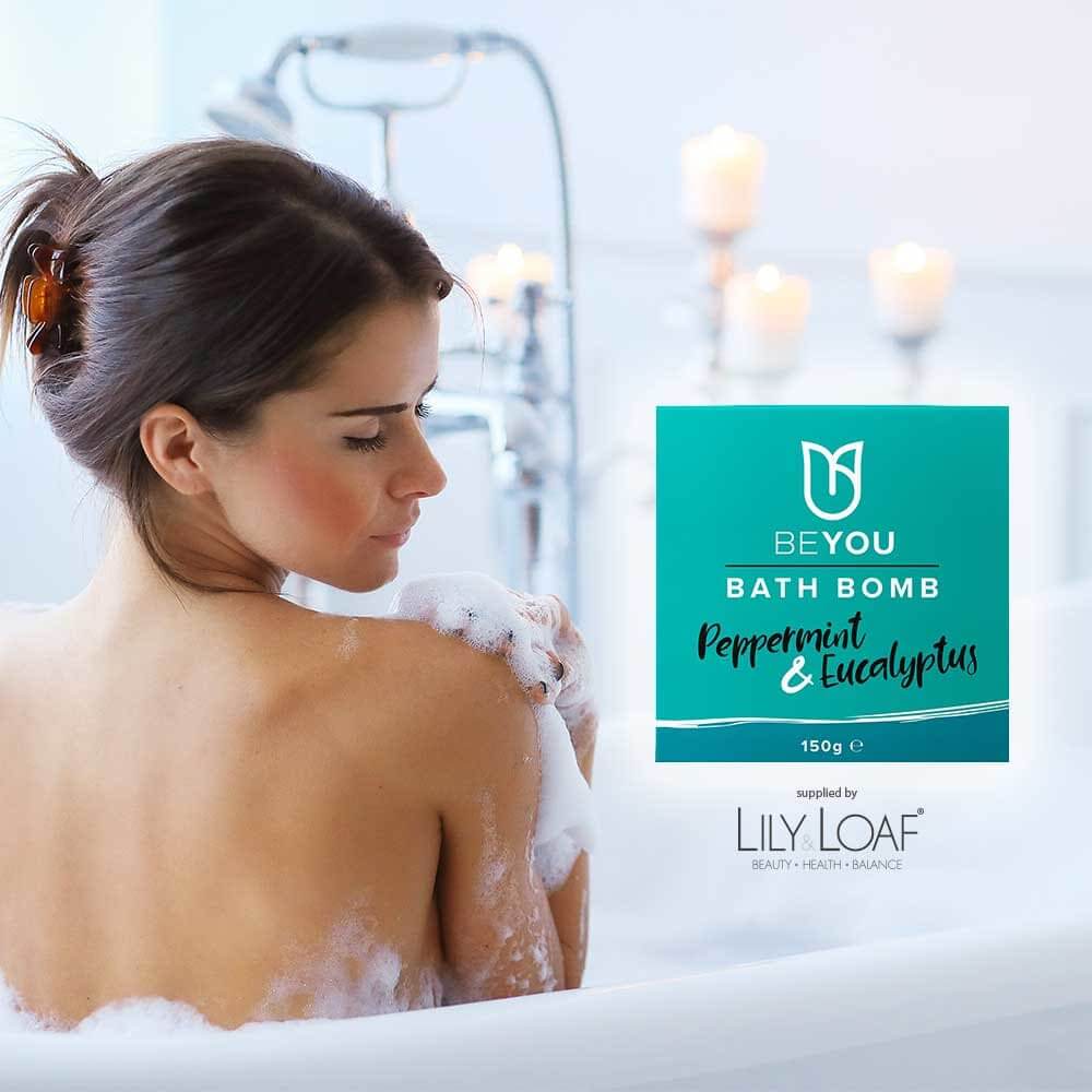 Woman relaxing in a bubble bath with candles, next to BeYou Peppermint & Eucalyptus Bath Bomb packaging, supplied by Lily & Loaf