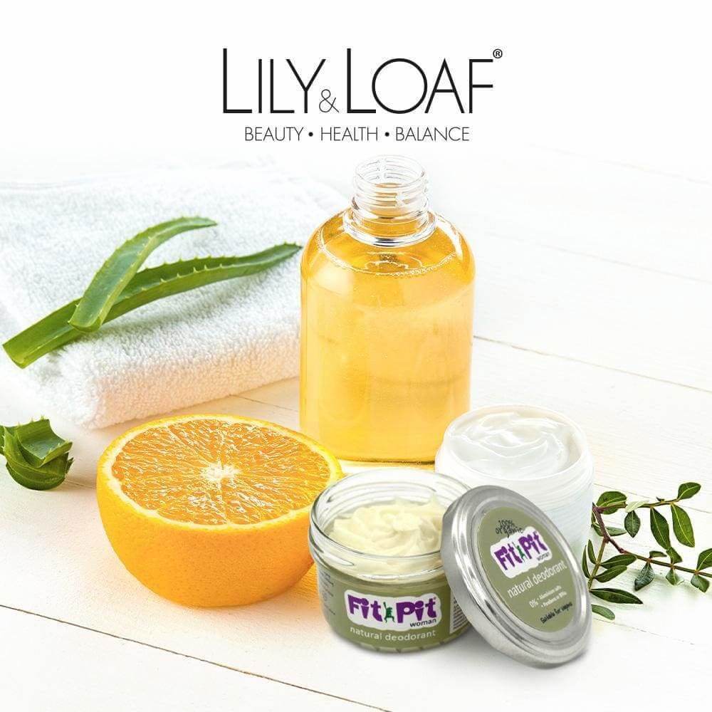 Fit Pit natural deodorant in small open glass jar with lid beside it, a bottle of golden liquid, half an orange, aloe vera leaf, white towel, white cream jar, and green leaves on a white wooden surface.