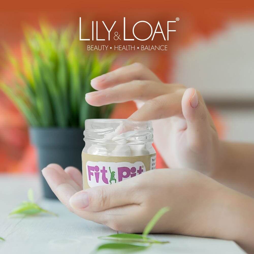 Hands scooping natural deodorant from open Fit Pit jar on table. Green plant and orange wall background. Lily & Loaf logo top centre.