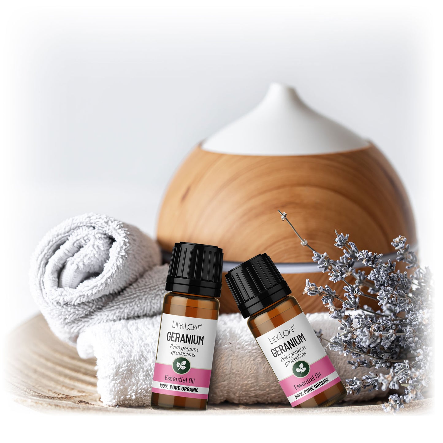 Two 10ml Geranium essential oil bottles with dried lavender, rolled towels, and a wooden diffuser on light wood, promoting wellness