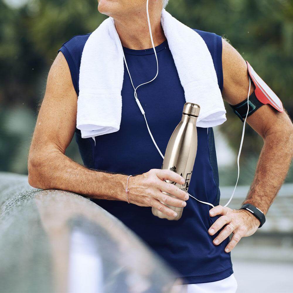 Fit older man wearing a navy sleeveless top and white towel around his neck, holding a stainless steel water bottle outdoors during exercise