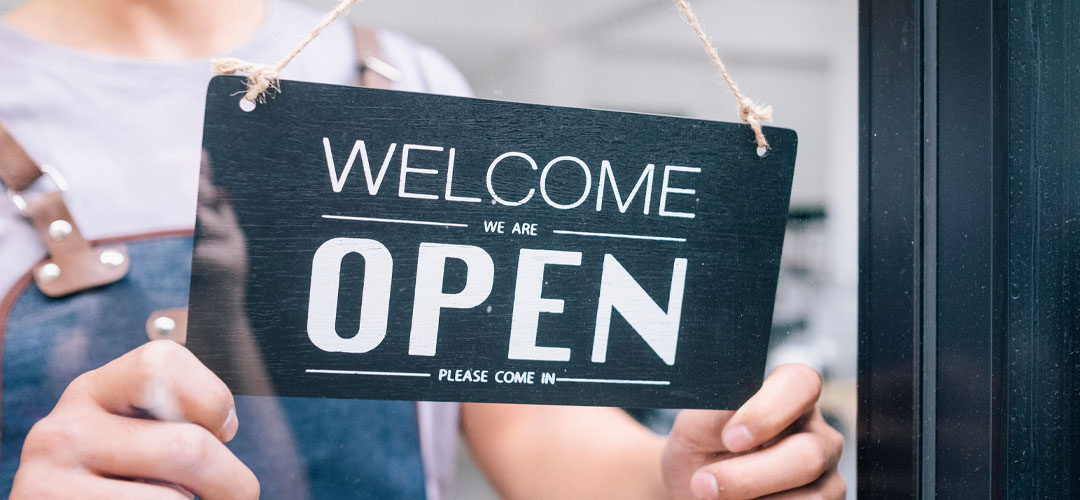 A person holding a black 'Welcome, we are open' sign at the entrance of a shop or business