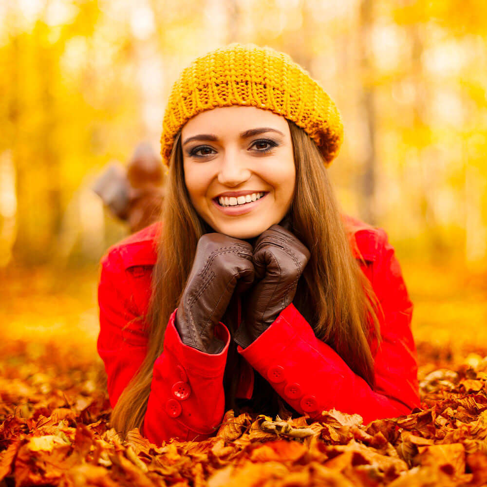 A woman in a yellow beanie and red coat smiling in autumn leaves.