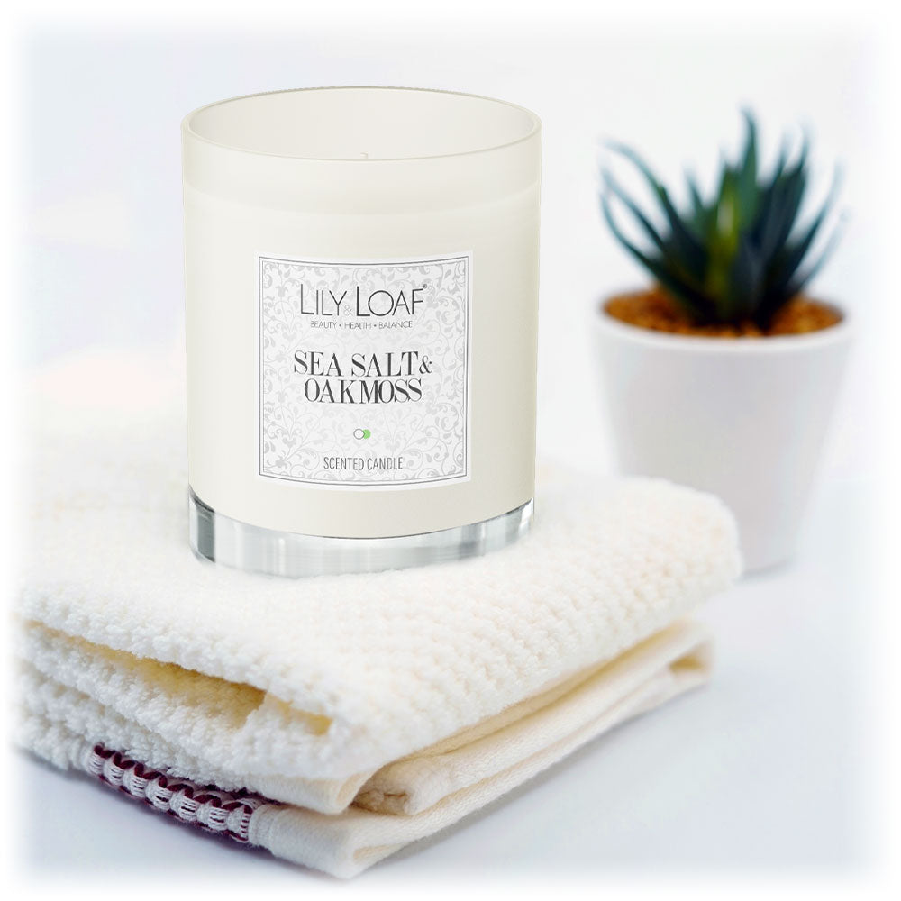 Lily & Loaf Sea Salt & Oakmoss Soy Wax Candle resting on a stack of fluffy white towels with a plant in the background